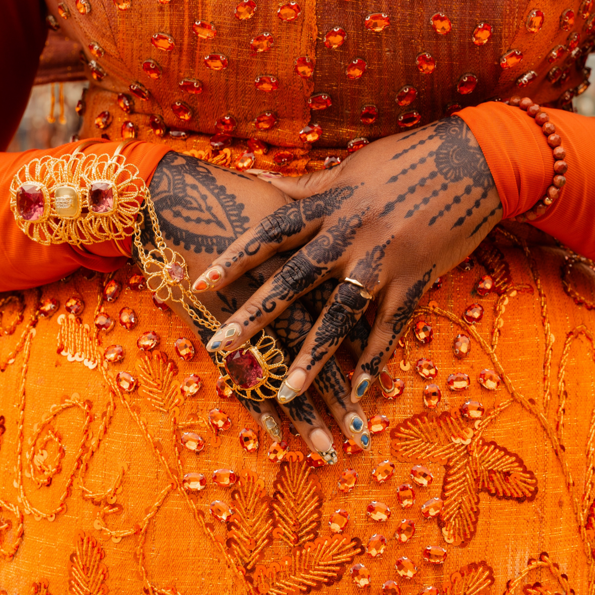 Henna-adorned hands with gold rings against an orange embroidered outfit, celebrating Yoruba culture