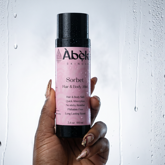 A hand with metallic gold manicured nails holds a bottle of Abeke Skincare Sorbet Hair & Body Mist against a glass door with water droplets.